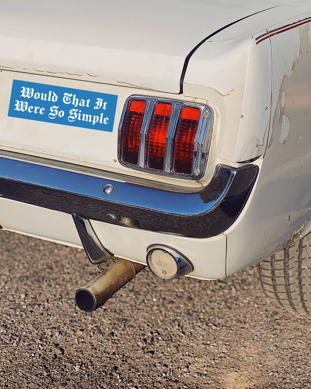 A close-up of the rear left side of a white classic car on gravel, showing a blue bumper sticker that reads &