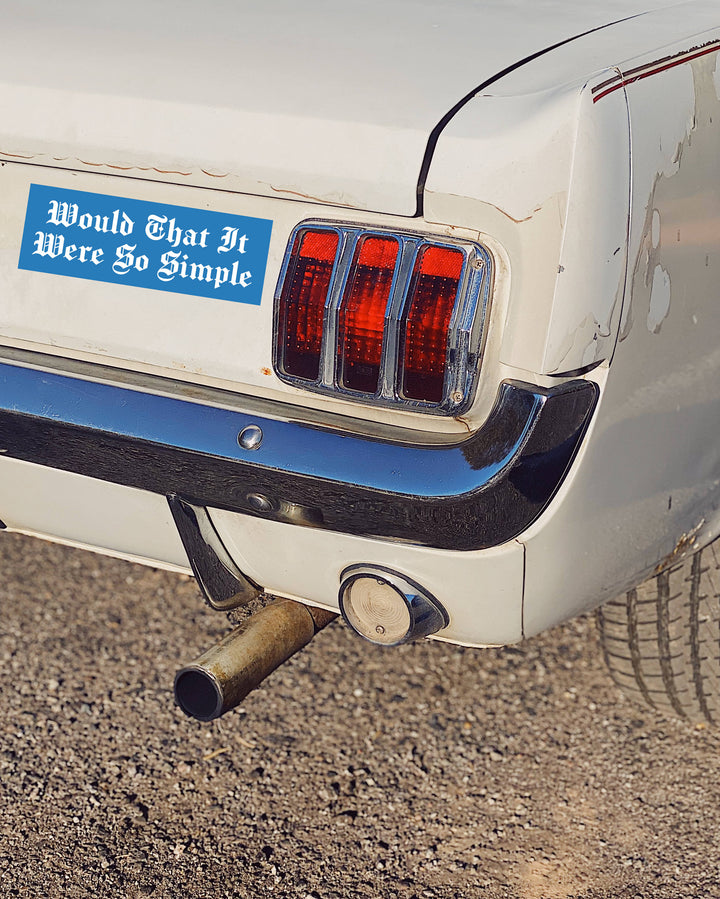 A close-up of the rear left side of a white classic car on gravel, showing a blue bumper sticker that reads &