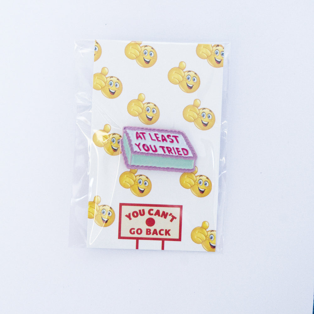 A novelty enamel lapel pin shaped like a motel sign with the text 'AT LEAST YOU TRIED' in pink on a light blue background, displayed on its white backing card with yellow emoji faces. A second sign pin reading 'YOU CAN'T GO BACK' is shown below.