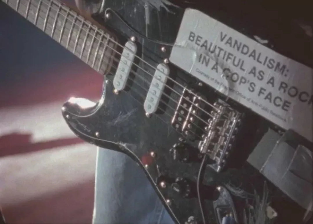 Guitar with a label reading 'Vandalism: Beautiful as a Rock in a Cop's Face' against a blurred background.